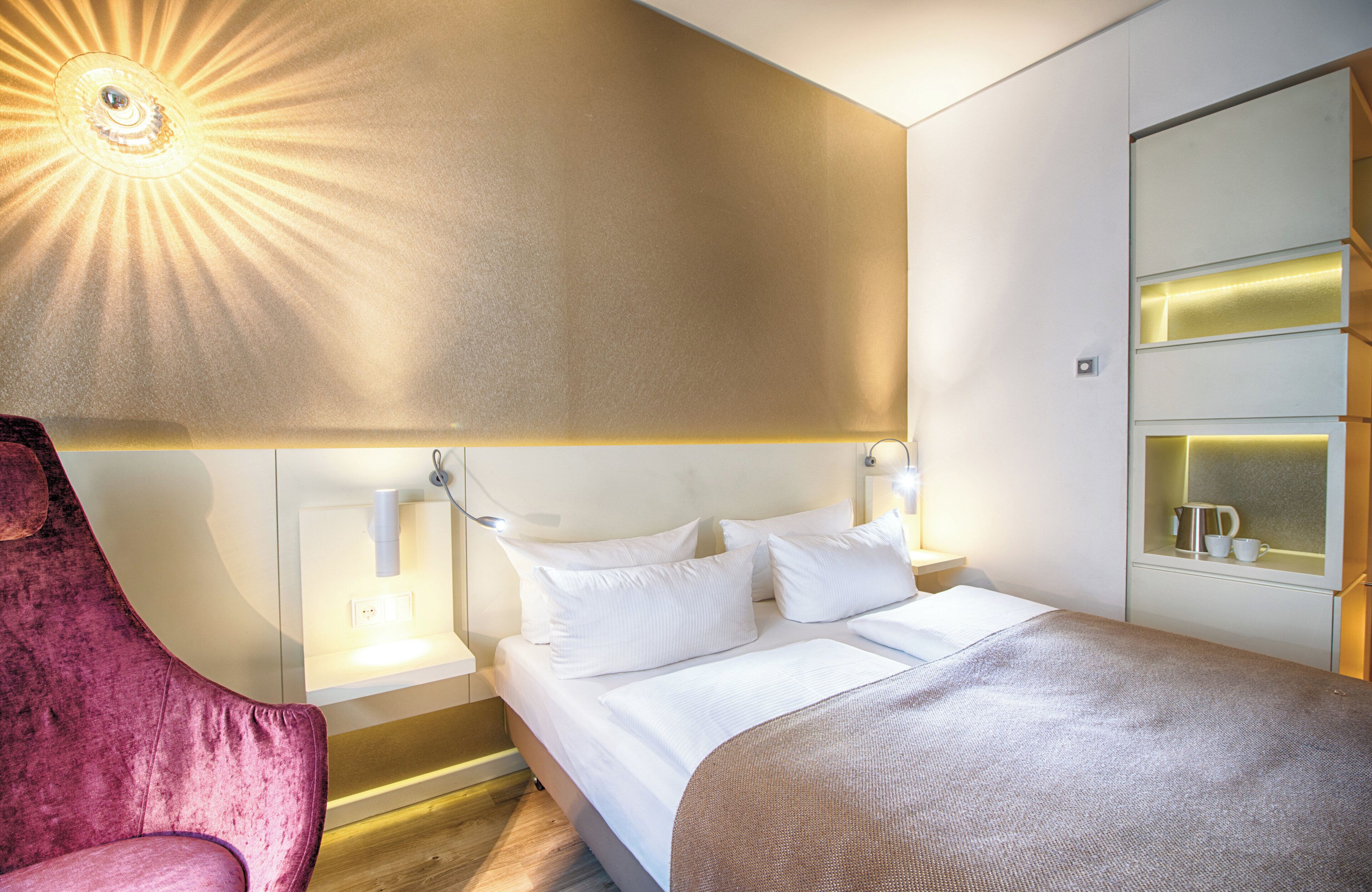 A modern hotel room with a large bed, a bedside table, a chair, and a wall-mounted shelf with a cup and a book.
