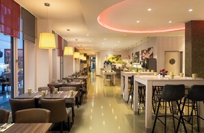 A modern hotel restaurant with multiple dining areas, a central bar, and a kitchen visible in the background. - Leonardo Hotel Berlin Mitte (Berlin)