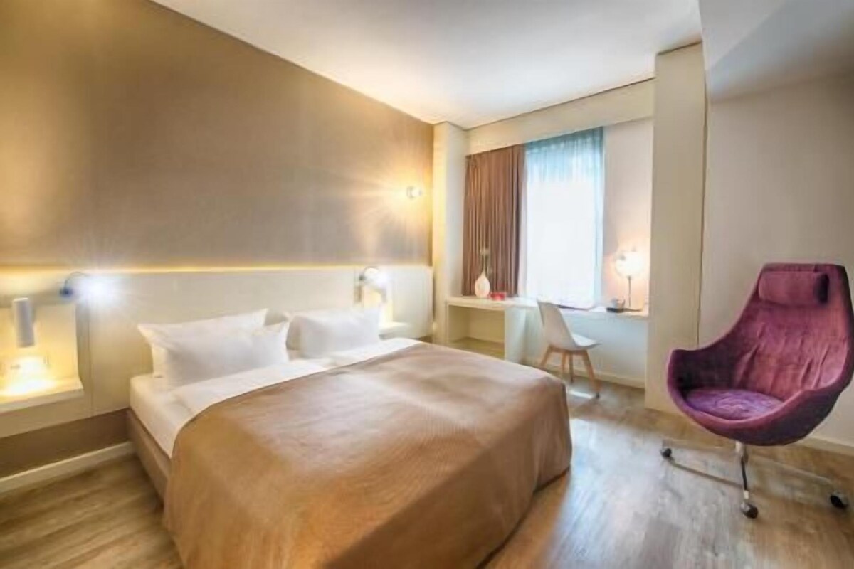 A hotel room with a bed, a desk, a chair, and a window with curtains.