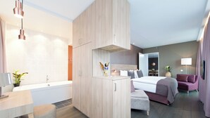 Habitación de hotel moderna con una cama grande, zona de estar separada, baño con bañera y escritorio con lámpara y planta.