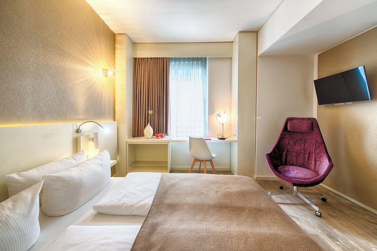 A hotel room with a bed, desk, chair, TV, and a window with curtains.