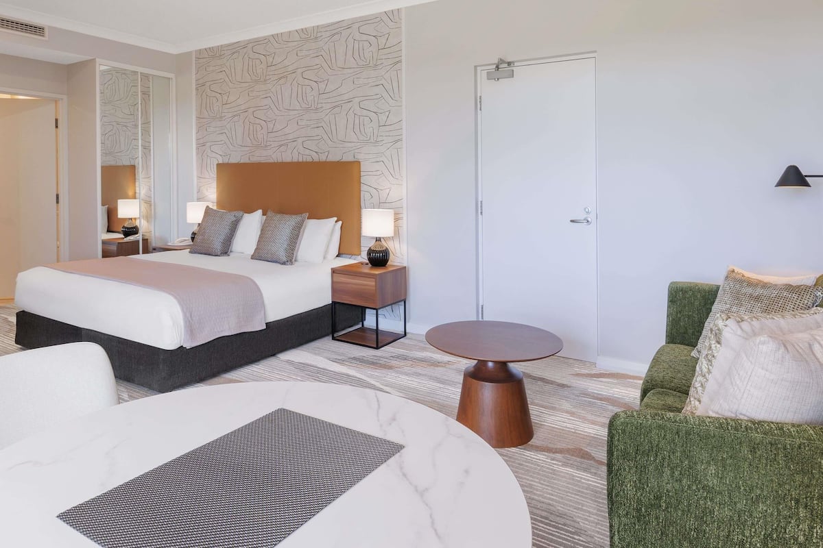 Ein Hotelzimmer mit einem großen Bett, einem grünen Sessel, einem runden Couchtisch und einem gemusterten Teppich.