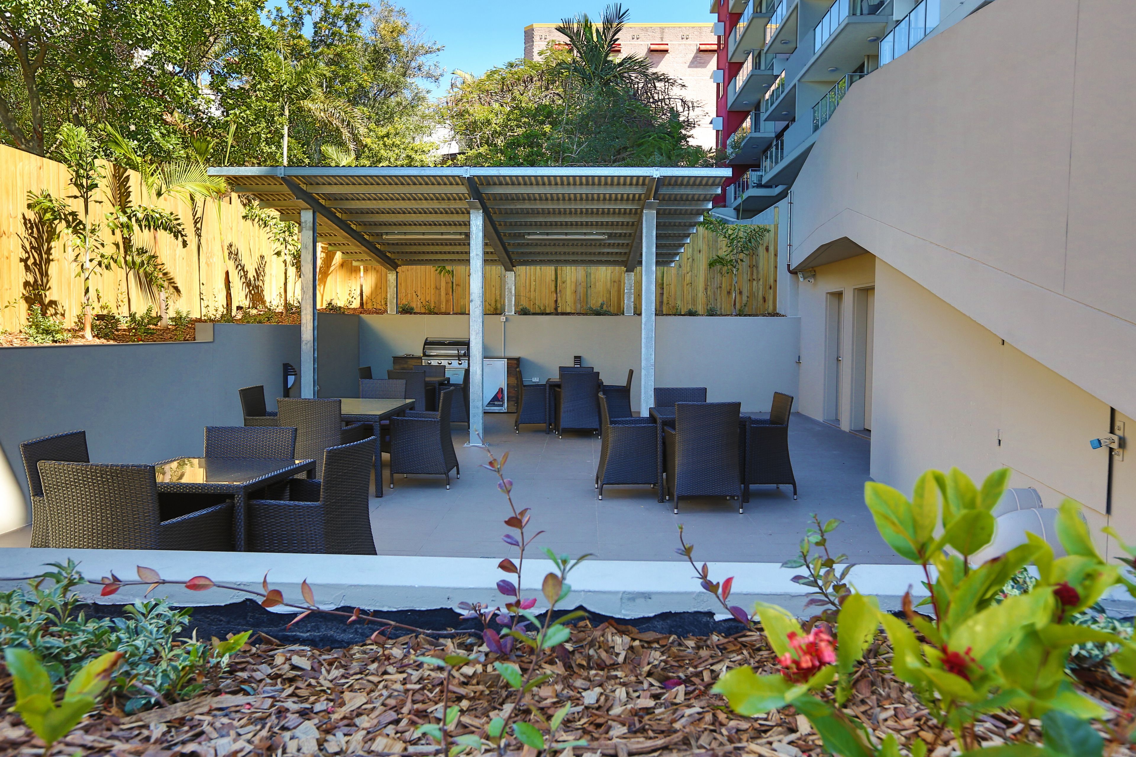 Un patio al aire libre con mobiliario de mimbre y un área de estar techada, rodeado de plantas y una valla de madera.