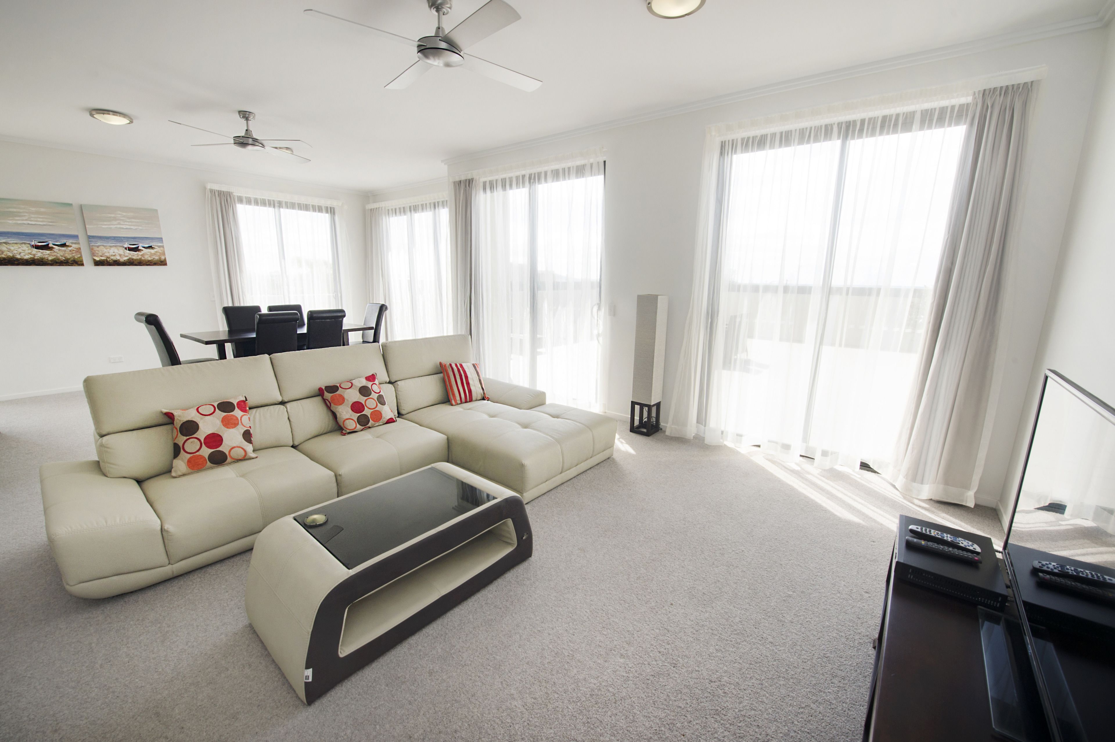 Una sala de estar moderna con un sofá seccional color beige, una mesa de centro de vidrio y un mueble para la televisión con controles remotos.