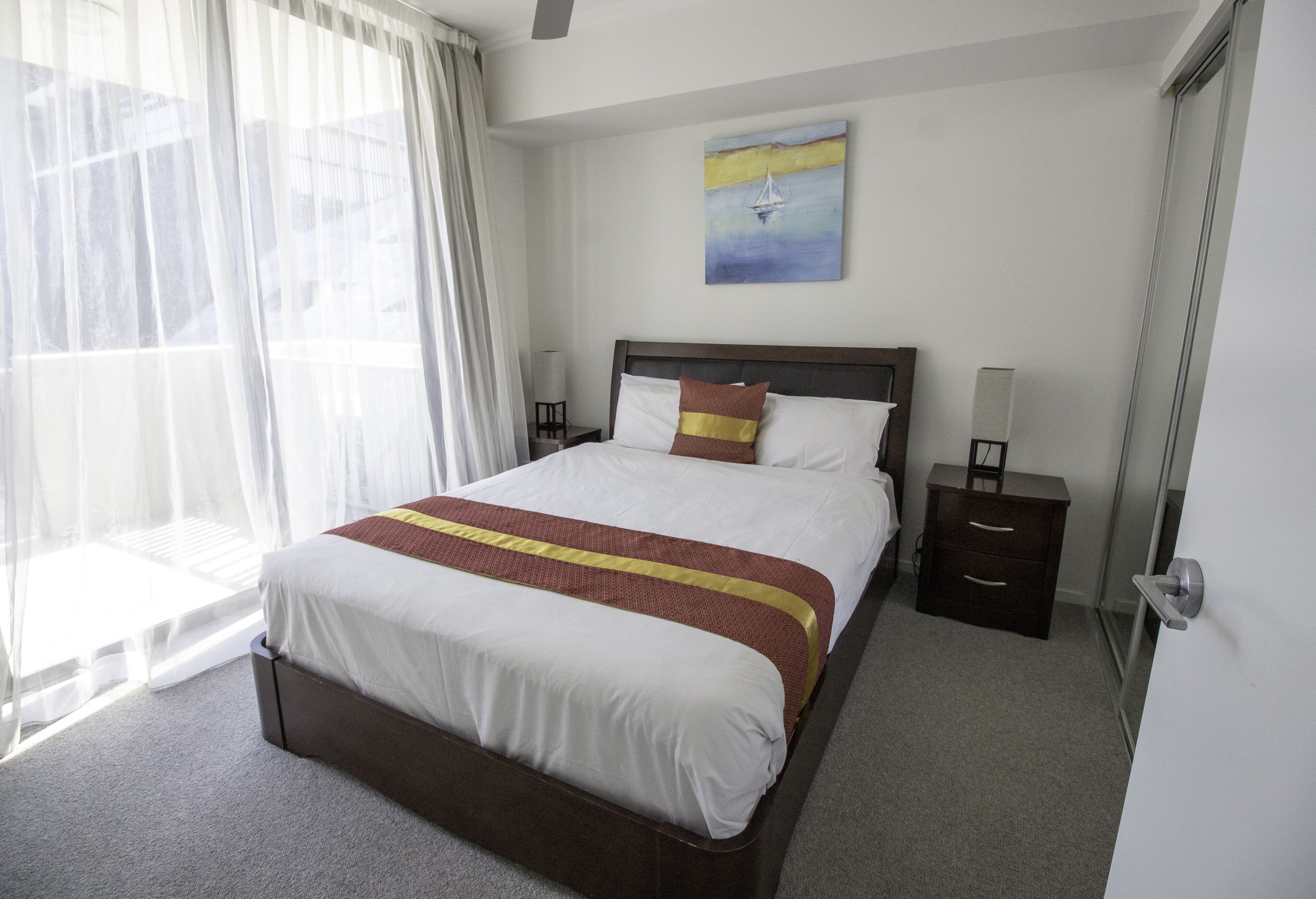 A neatly made bed with a brown headboard, a bedside table with a lamp, and a painting on the wall.