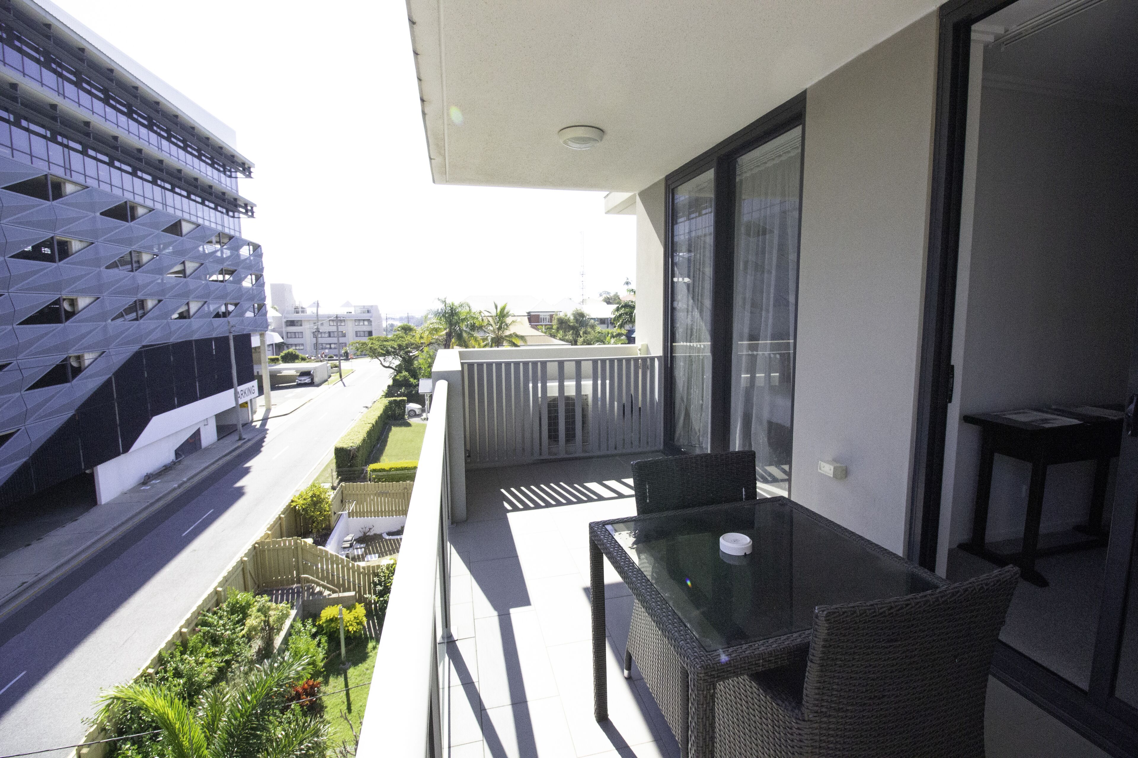 Un balcon doté d’une table et de chaises en verre, donnant sur une rue bordée d’immeubles et d’espaces verts.