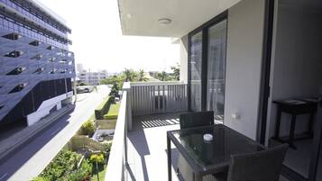 Un balcon doté d’une table et de chaises en verre, donnant sur une rue bordée d’immeubles et d’espaces verts.