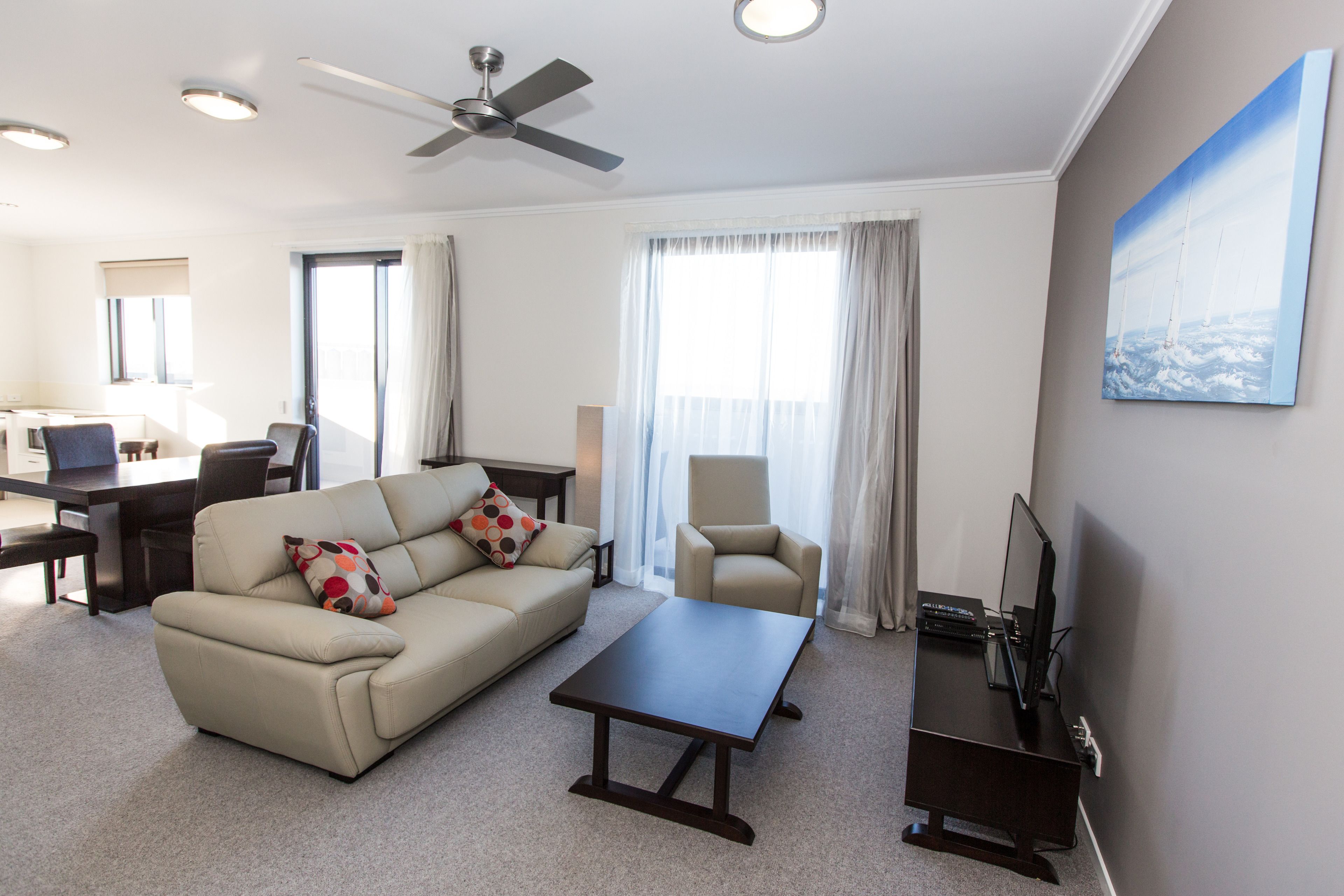 A modern hotel room with a sofa, armchair, coffee table, desk, and a flat-screen TV.