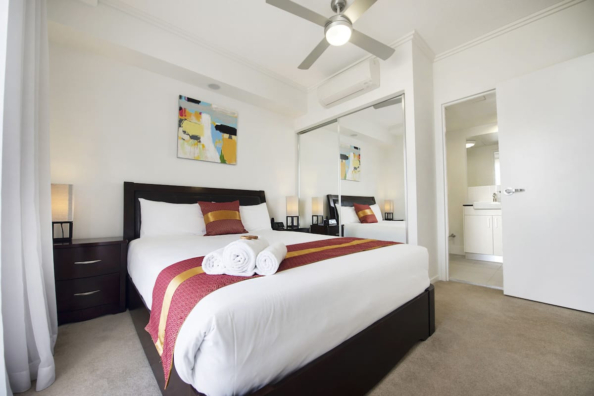 Een hotelkamer met een groot bed, een plafondventilator, een nachtkastje met een lamp, een spiegel en uitzicht op de badkamer.