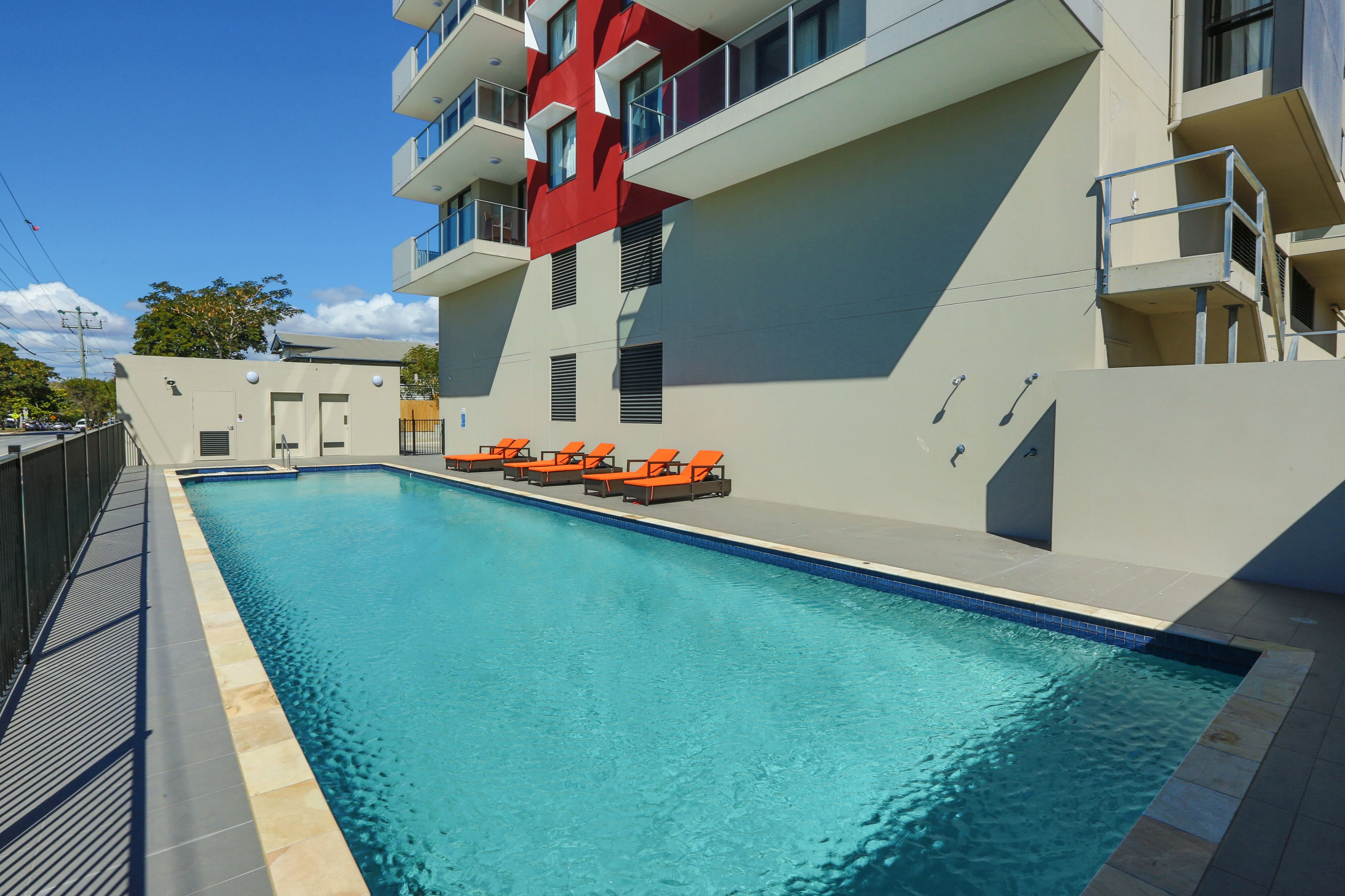 A swimming pool with orange lounge chairs, a modern building with balconies, and a clear blue sky.
