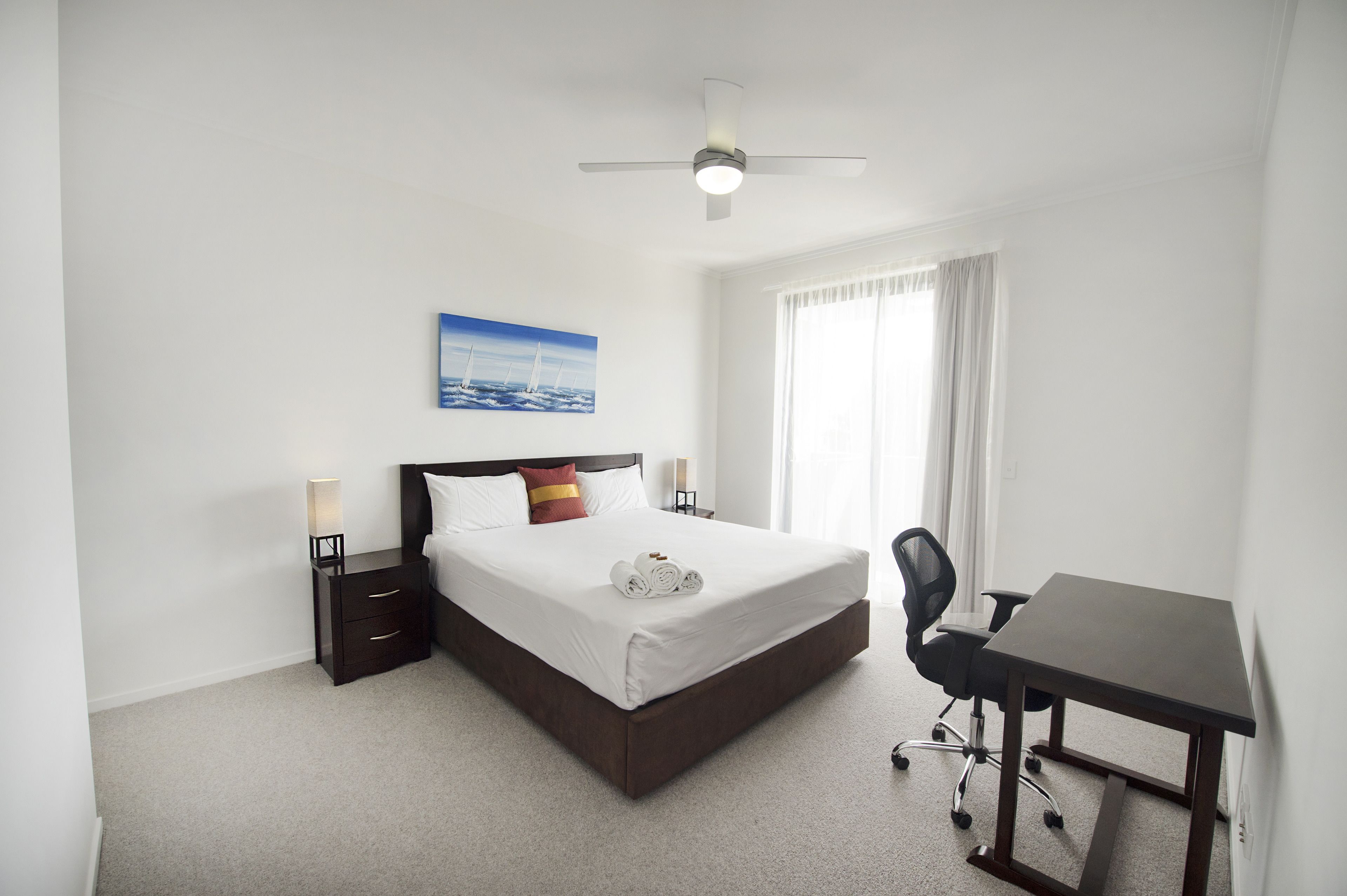 Une chambre bien rangée, avec un lit, des lampes de chevet, un bureau et un ventilateur de plafond.