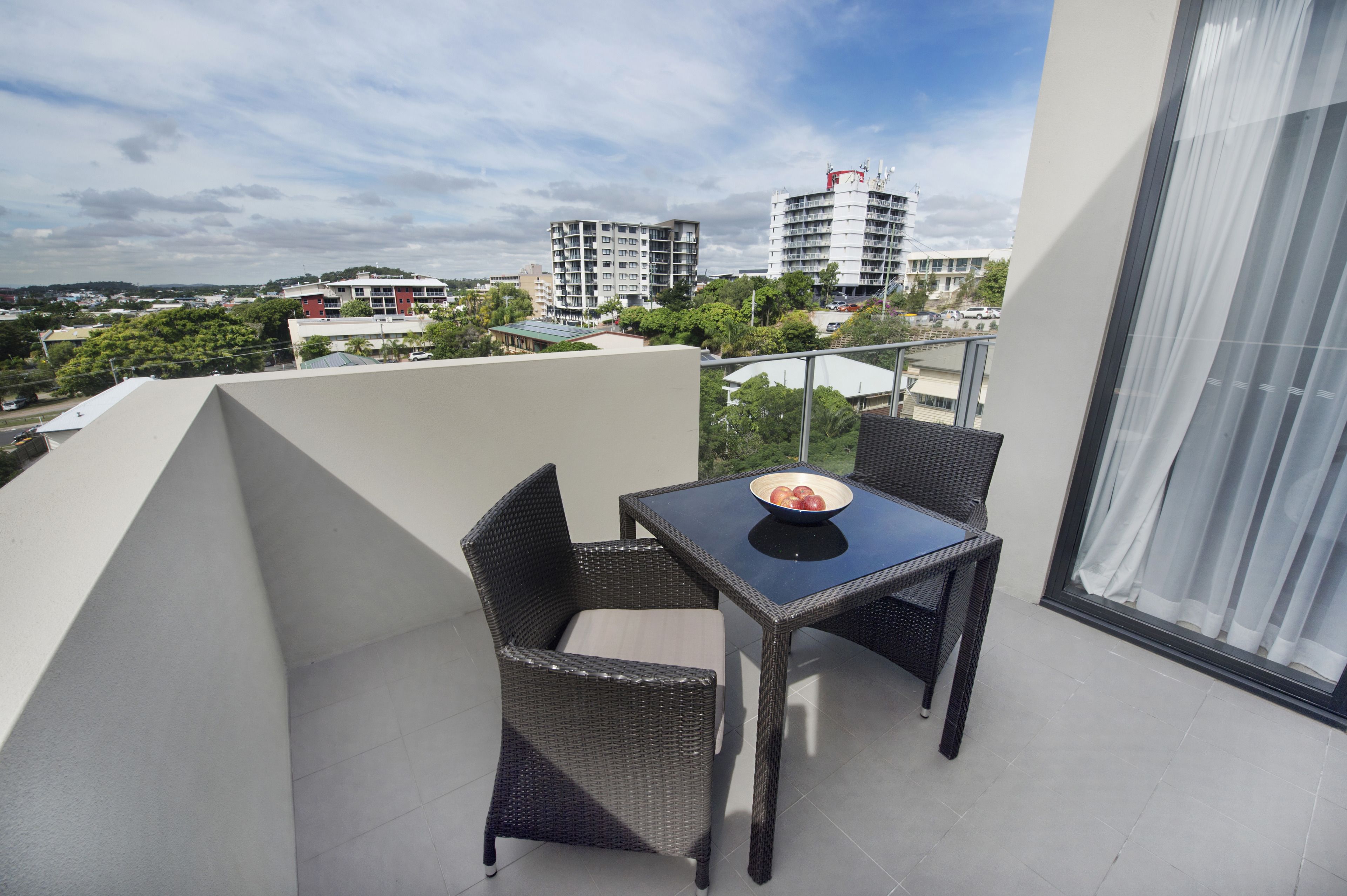 Ein Balkon mit Korbmöbeln und einem Tisch, mit Blick auf eine Stadtlandschaft aus Gebäuden und Grünflächen.