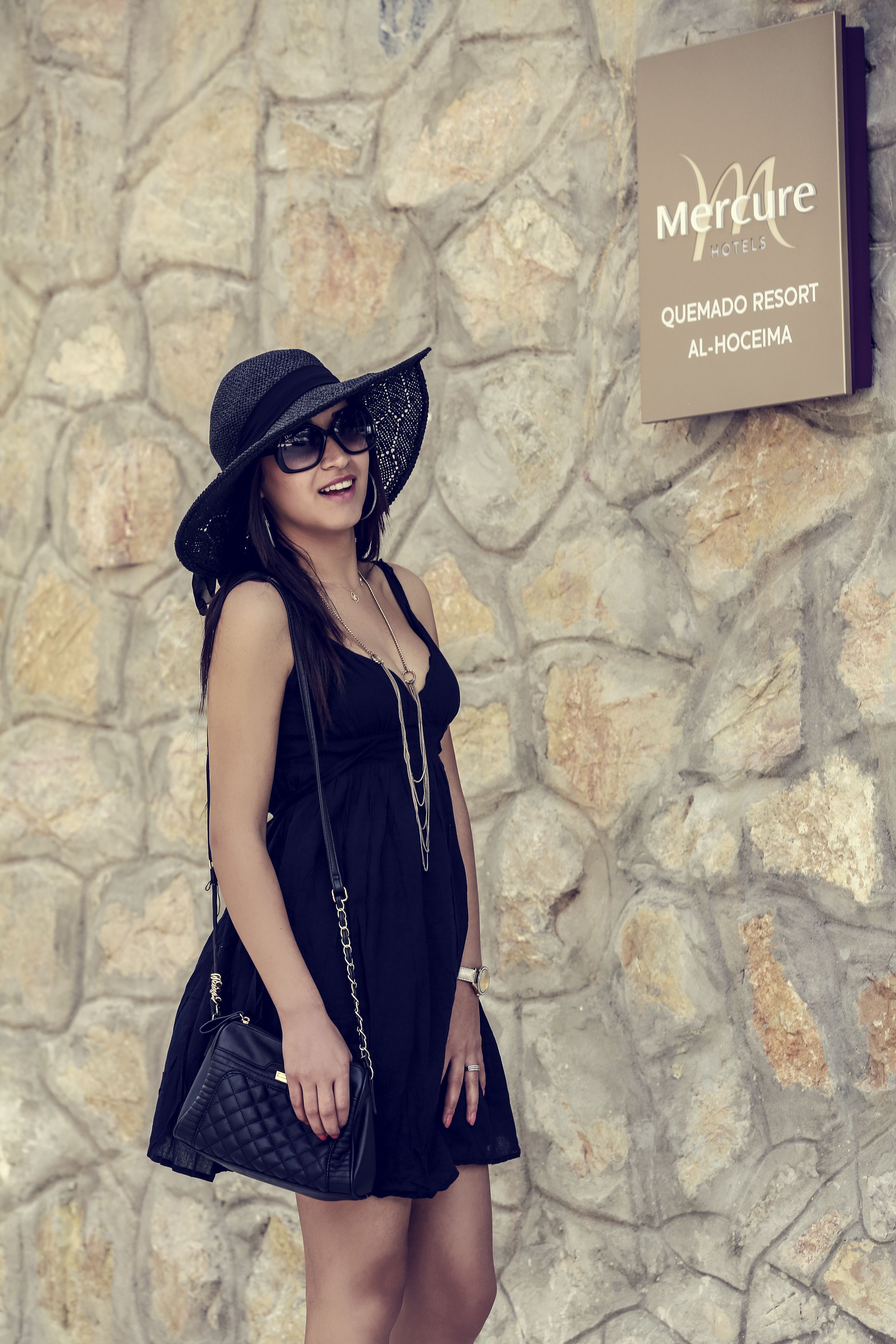 A woman in a black dress and hat standing in front of a stone wall with a sign for Mercure Hotels.
