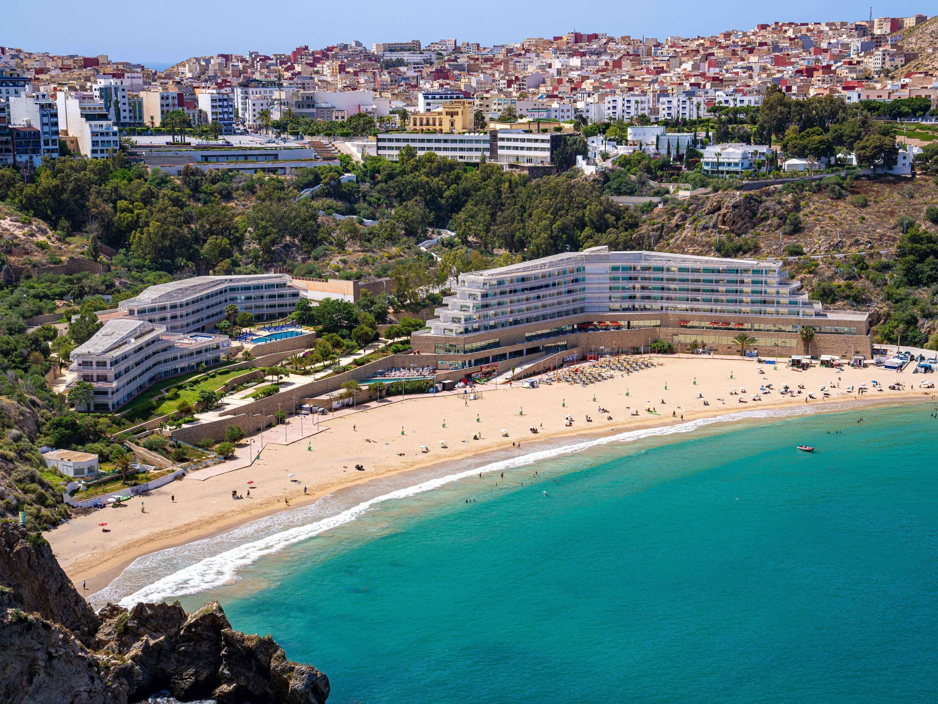 A coastal hotel with multiple buildings, a beach, and a view of the city in the background.
