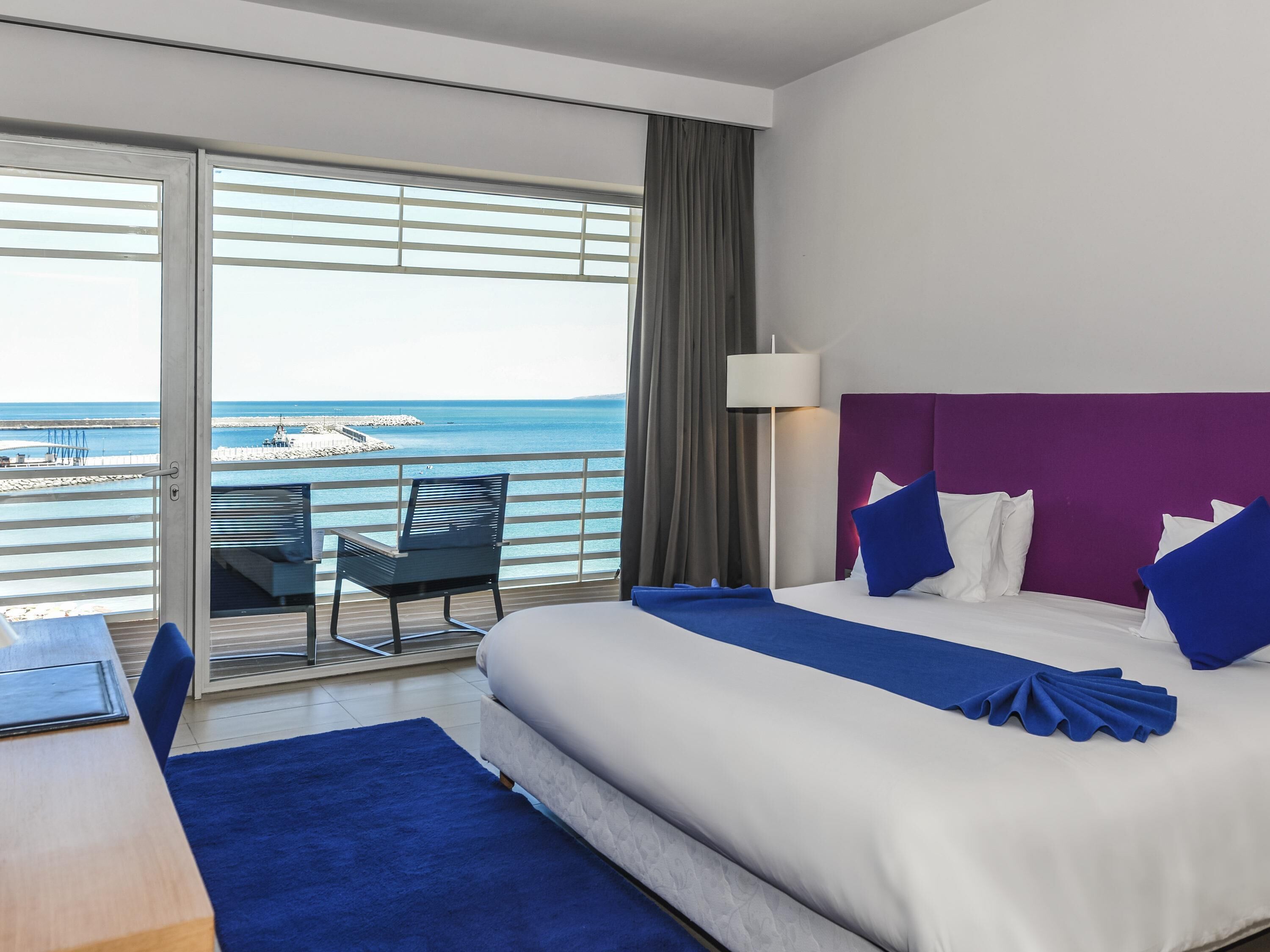 A hotel room with a bed, a desk, a chair, and a view of the sea.