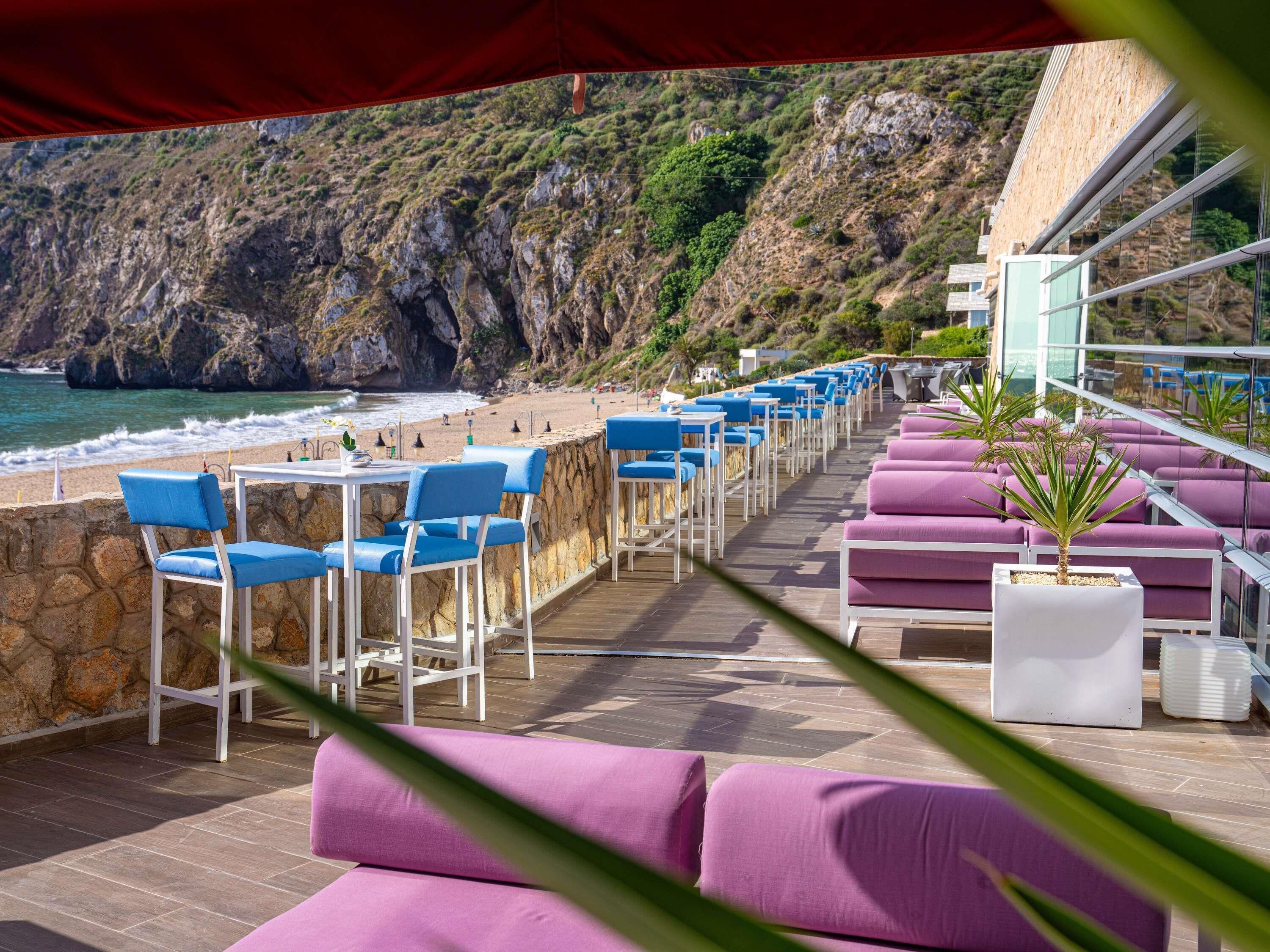 Eine Strandterrasse mit blau-violetten Sitzgelegenheiten, einer Steinsäule und Blick auf das Meer und die Klippen.