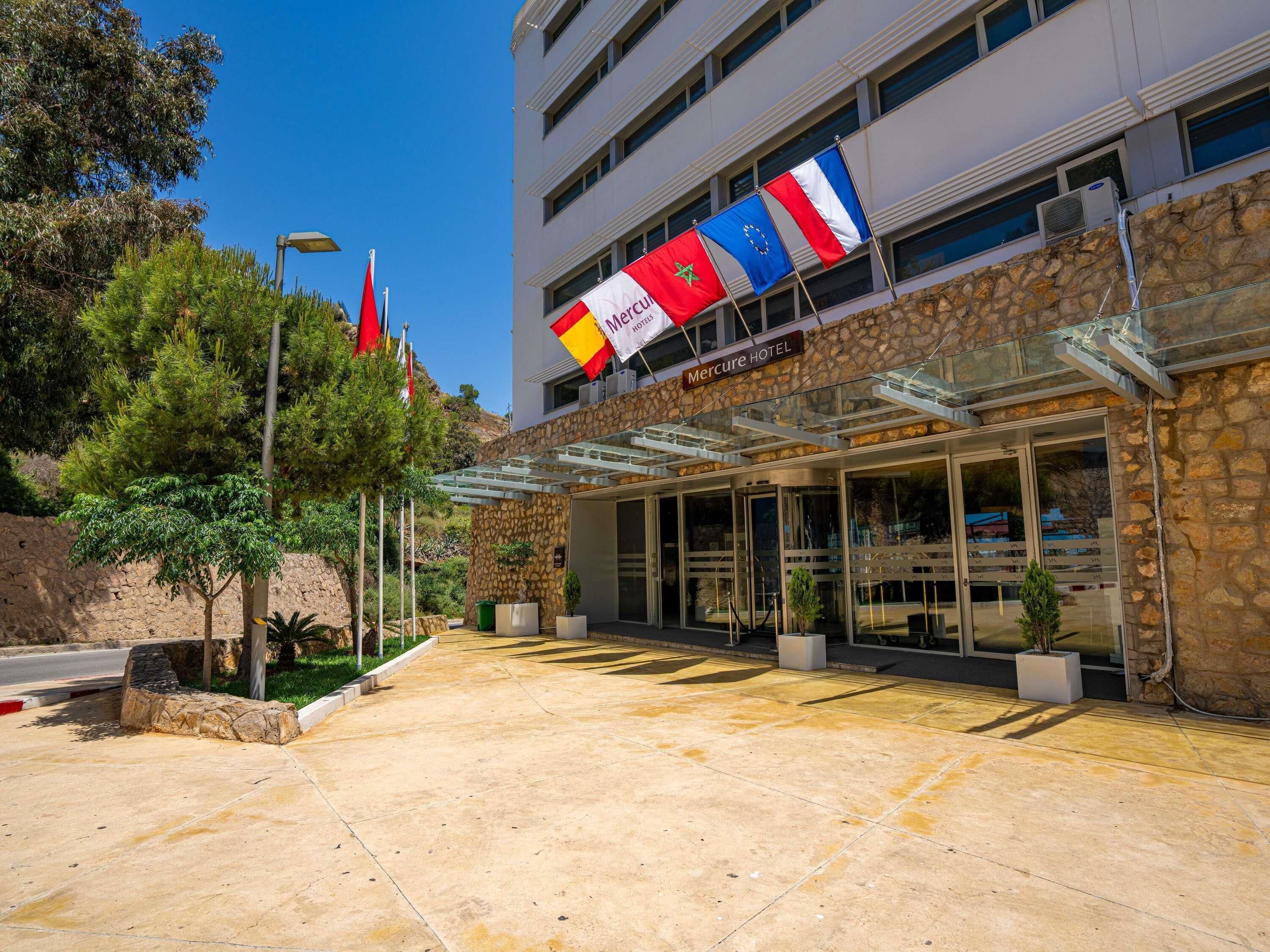 L’entrée d’un hôtel, avec une façade en pierre, des portes en verre et plusieurs drapeaux.