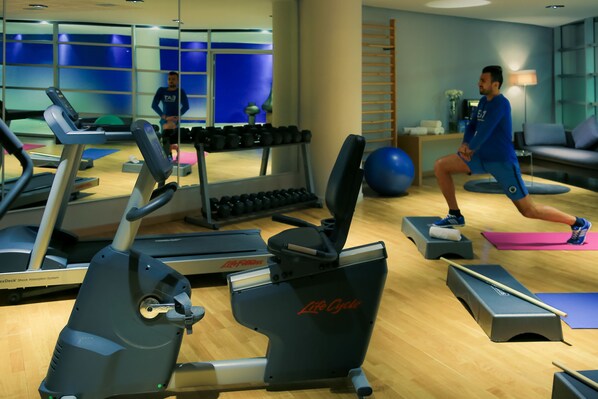 Un gimnasio con equipo cardiovascular, mancuernas y una persona usando una plataforma elevada.