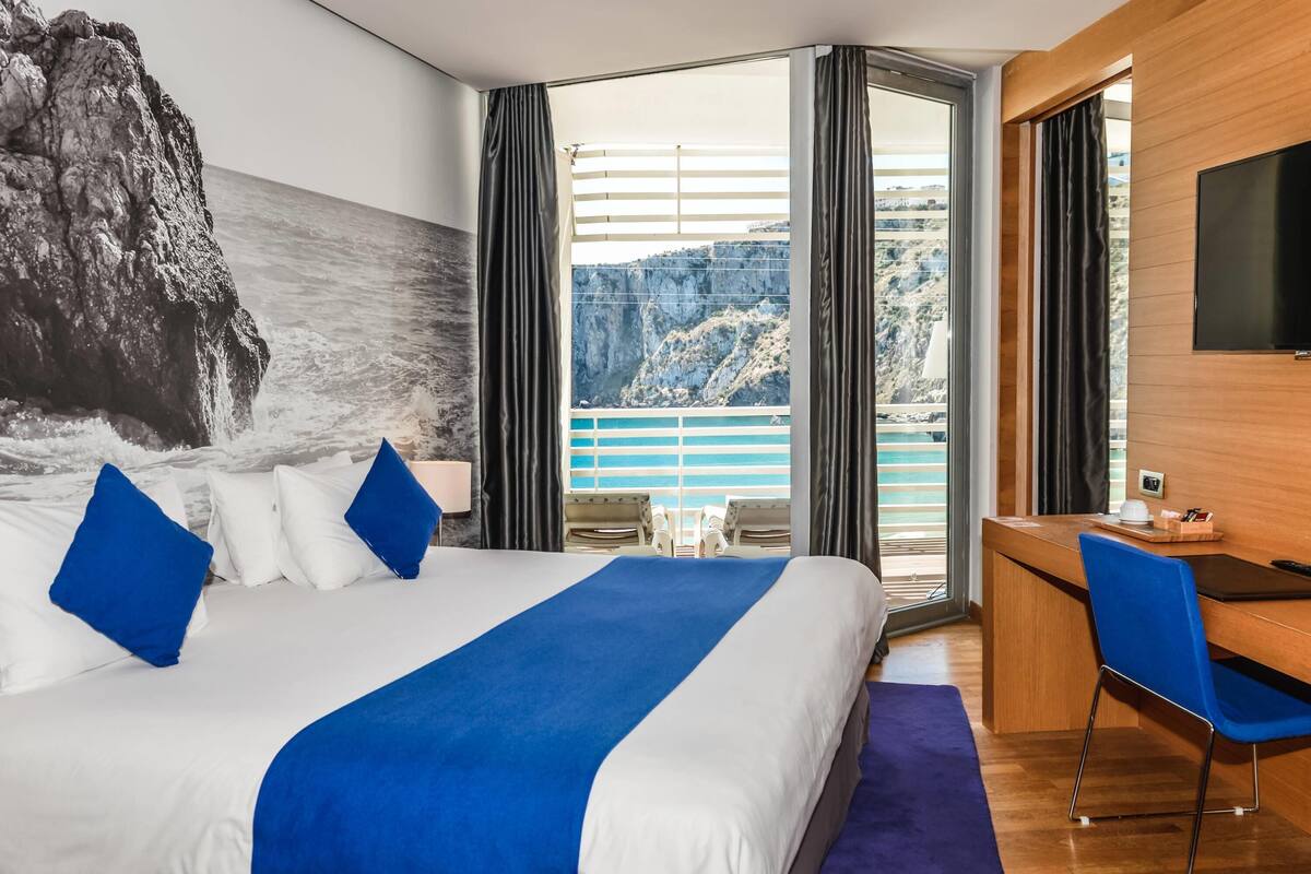 A hotel room with a bed, a desk, a chair, a TV, and a view of the sea and cliffs.