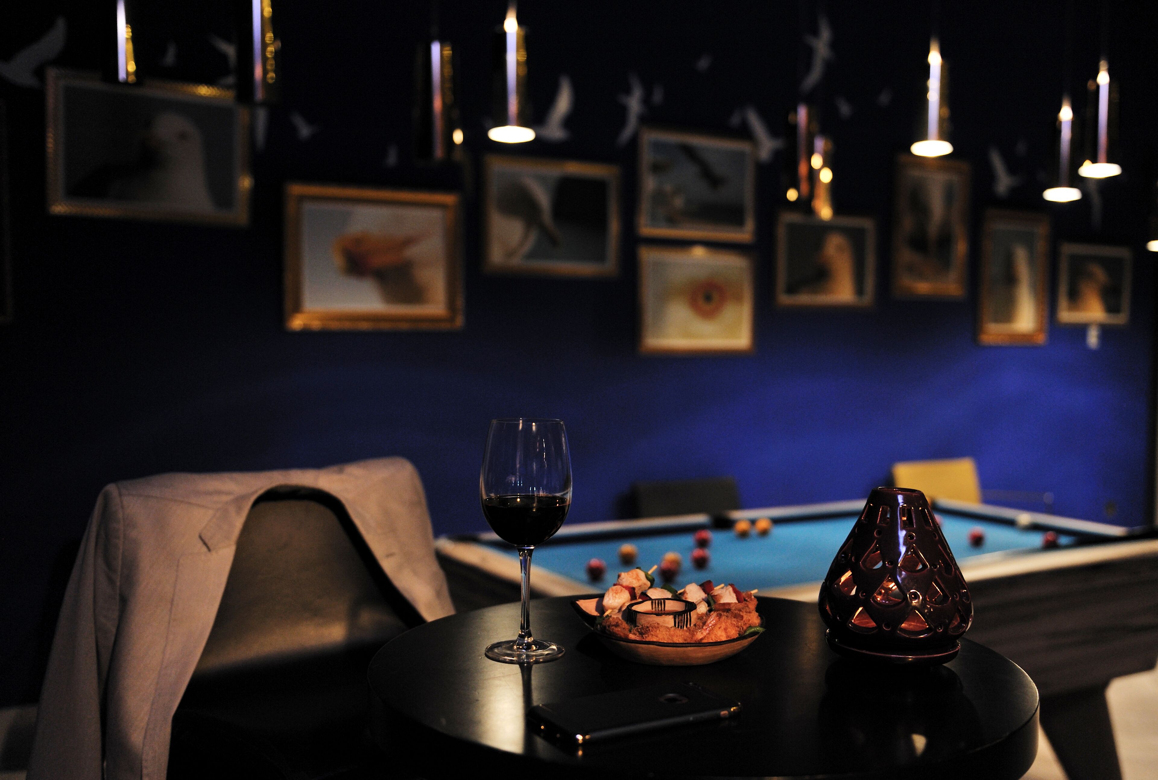 Une pièce faiblement éclairée, avec une table de billard, des tableaux encadrés accrochés au mur, un verre de vin rouge et un bol de collations posé sur une table.
