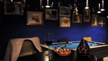 A dimly lit room with a pool table, framed pictures on the wall, a glass of red wine, and a bowl of snacks on a table.