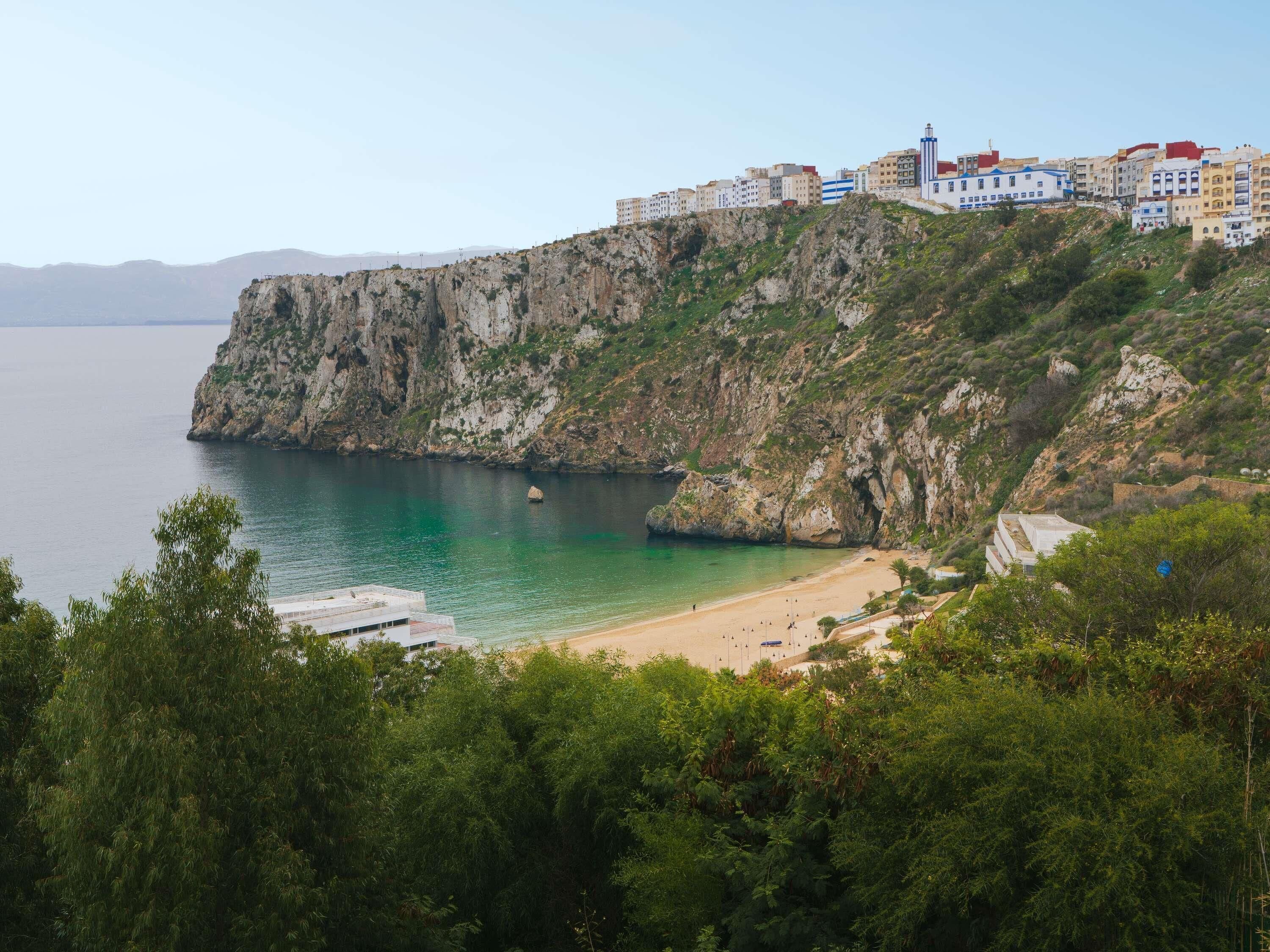 A coastal town with buildings on a cliff overlooking the sea, a beach, and dense greenery in the foreground.