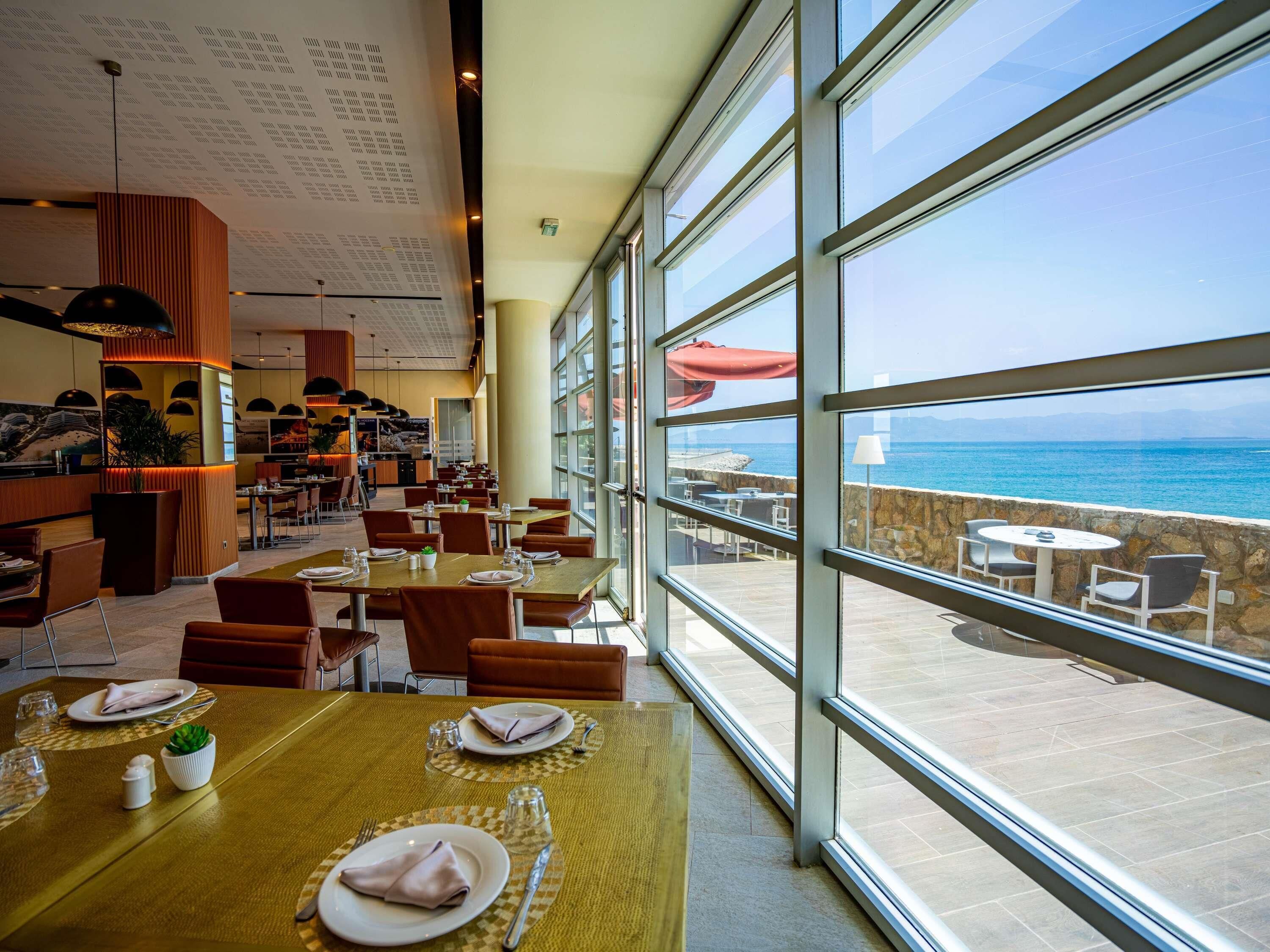 A restaurant with tables set for dining, a bar area, and a view of the sea through large windows.