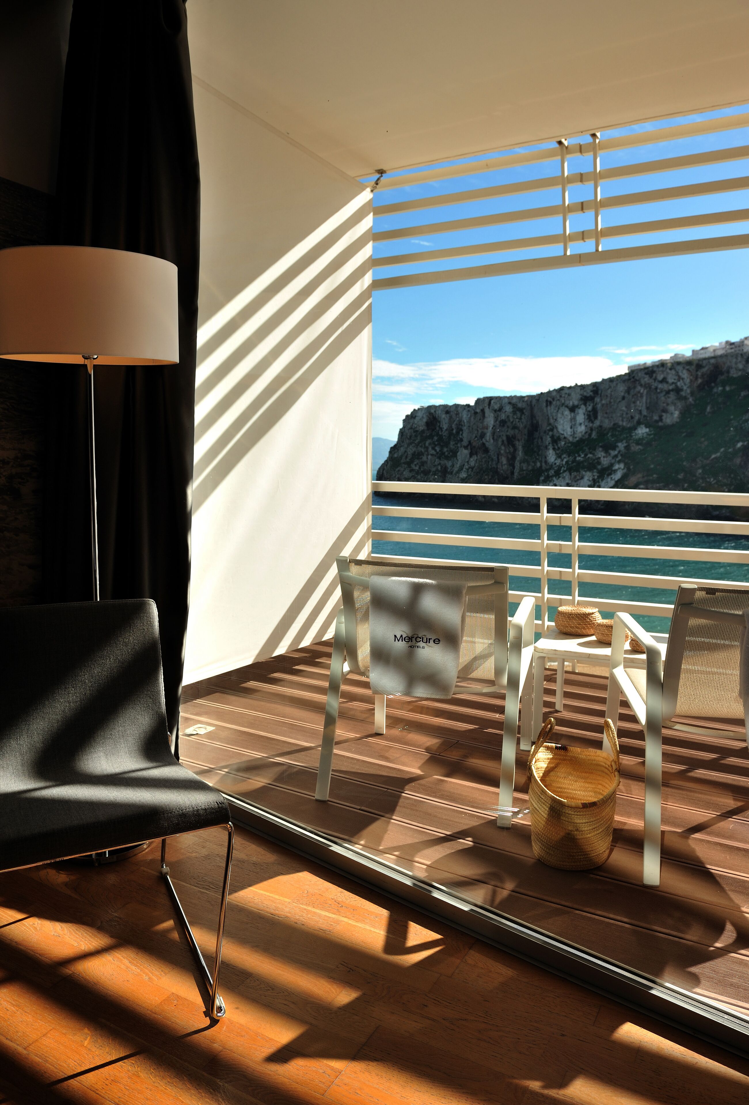 Varanda com vista para o mar e uma montanha, mobiliada com cadeiras e uma mesa, e um candeeiro.
