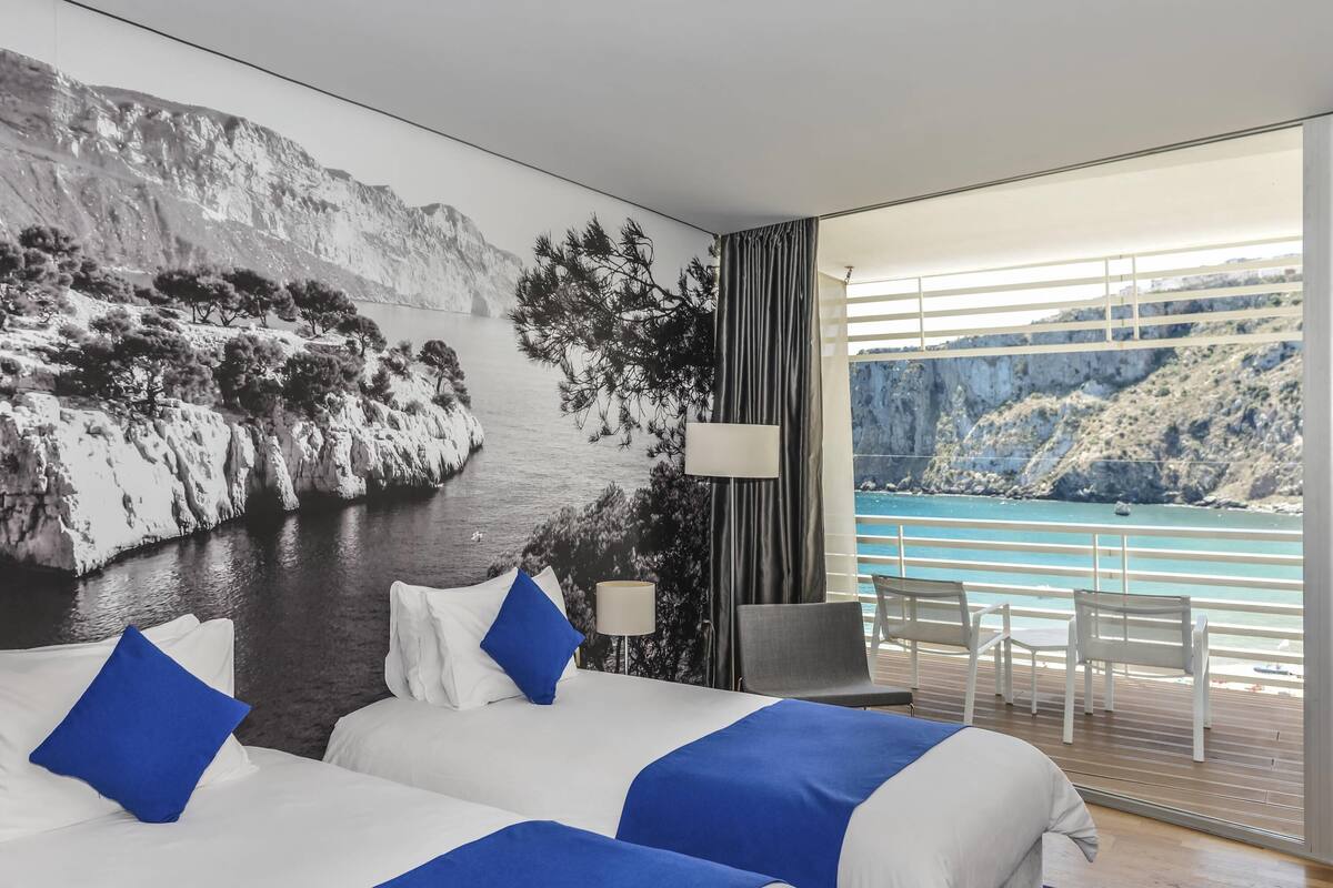 A hotel room with two beds, a view of a coastal landscape, a balcony with a table and chairs, and a large window.