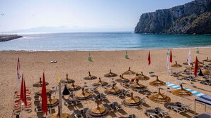 A beach with rows of sun loungers and umbrellas, a rocky cliff, and a clear sky. - Mercure Quemado Resort (Al Hoceima)
