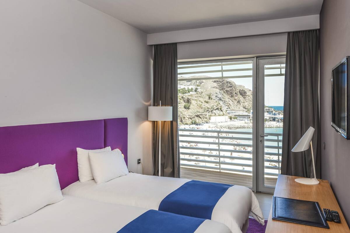 A hotel room with a bed, a desk, a lamp, and a view of a beach and mountains.