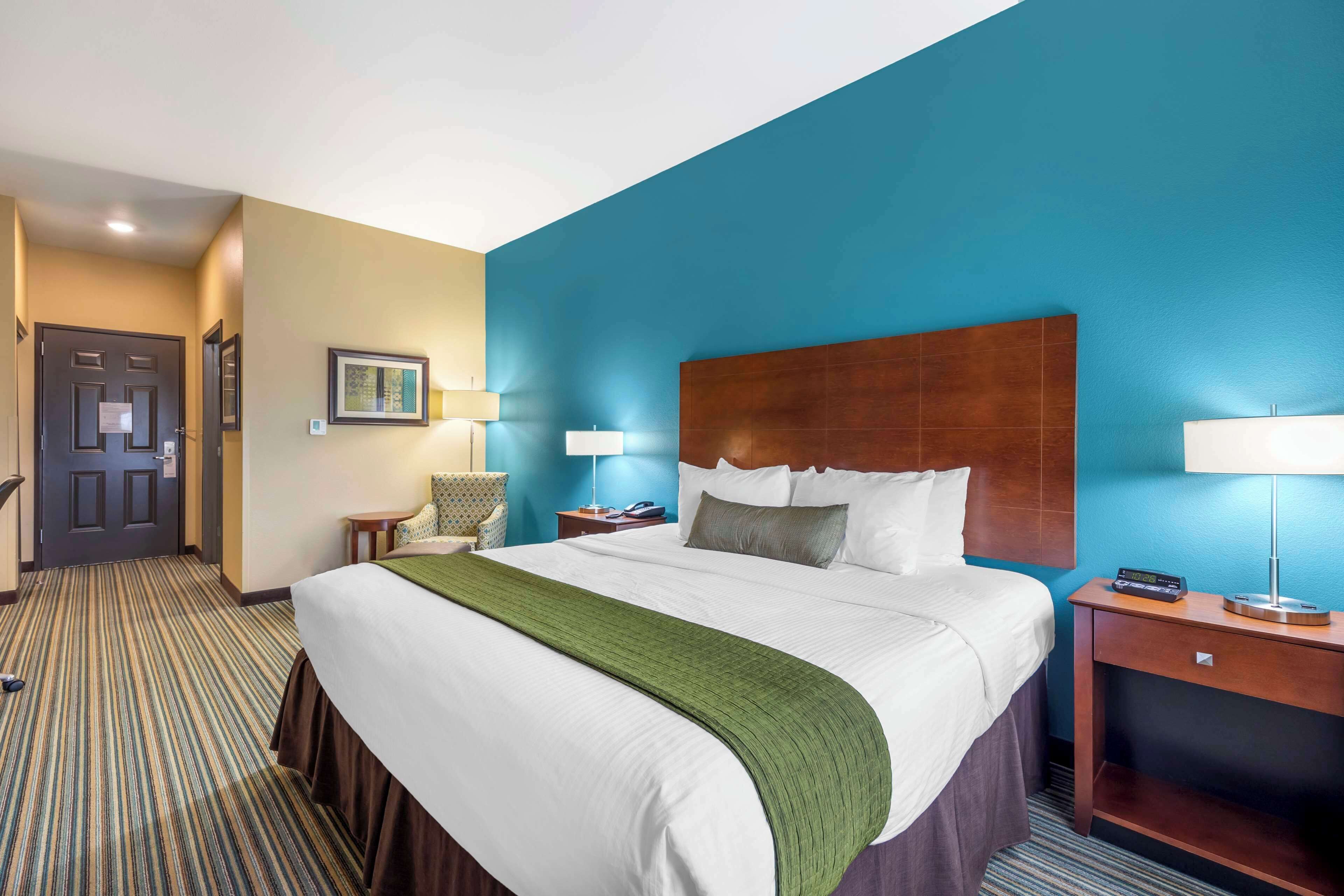 A hotel room with a large bed, a wooden headboard, two bedside tables with lamps, a green throw blanket, and a door with a sign.