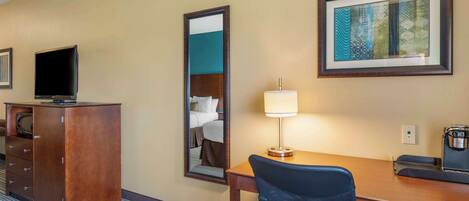 A hotel room with a wooden desk, a chair, a television, a mirror, a lamp, and a coffee maker.