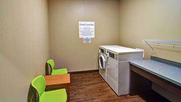 A small laundry area with a washing machine, a drying rack, and two green chairs.