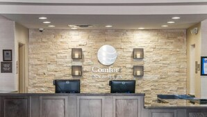 Balcão de recepção com parede de pedra ao fundo, dois monitores e uma placa com a inscrição 'Comfort Inn & Suites'.