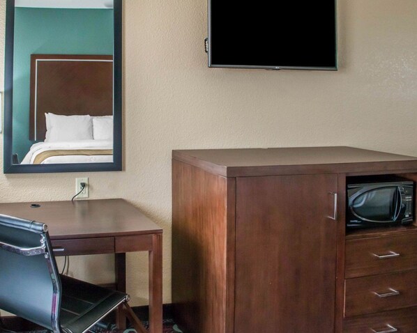 A hotel room with a wooden cabinet containing a microwave, a desk with a chair, and a mirror reflecting a bed.