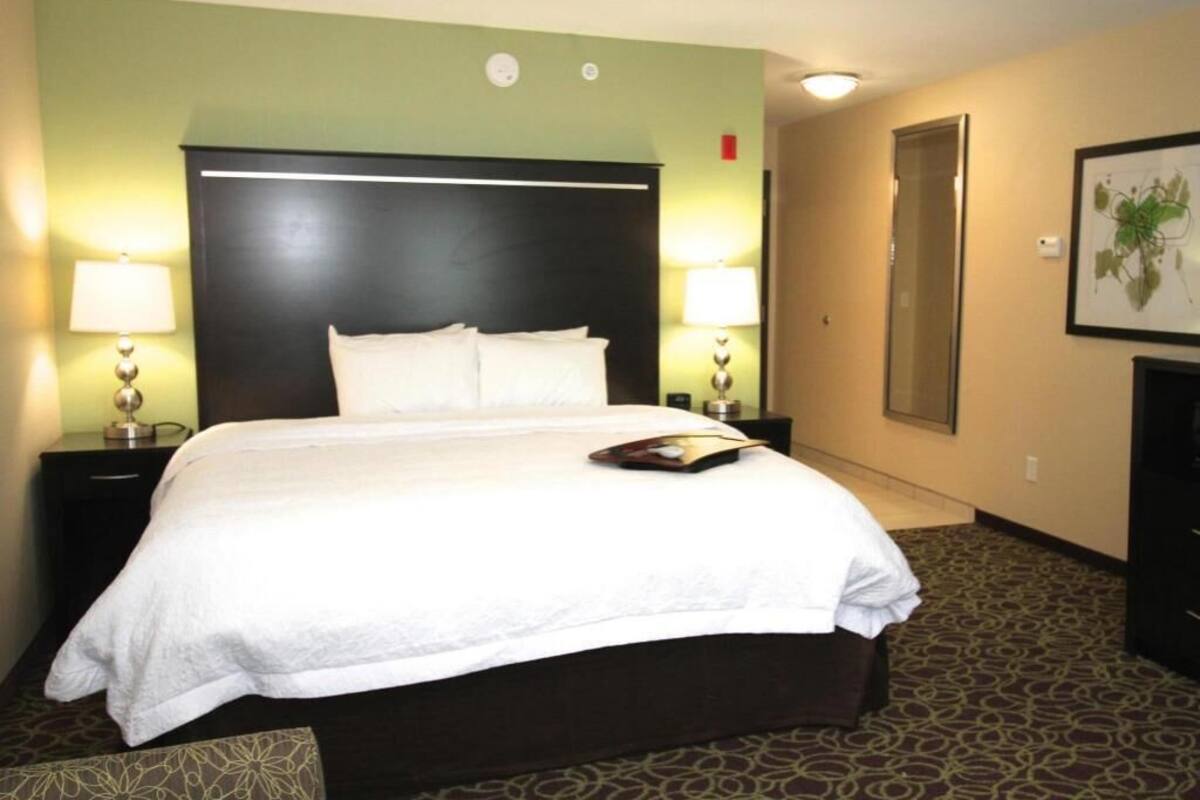 A hotel room with a large bed, two bedside tables with lamps, a headboard, a carpeted floor, and a framed picture on the wall.