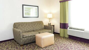 A hotel room with a patterned sofa, a small ottoman, a framed picture on the wall, a lamp, and an air conditioning unit.