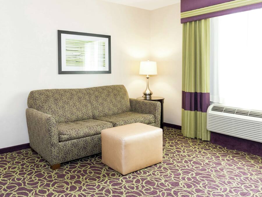A hotel room with a patterned sofa, a small ottoman, a framed picture on the wall, a lamp, and an air conditioning unit.