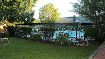 A swimming pool area with a covered seating area, surrounded by greenery and a brick pathway.