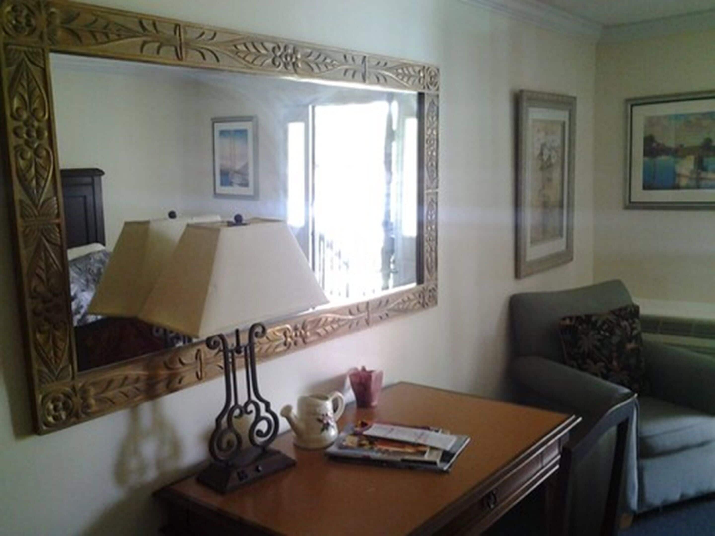 A room with a large ornate mirror, a lamp, a small table with a teapot and a magazine, a sofa, and framed pictures on the wall.