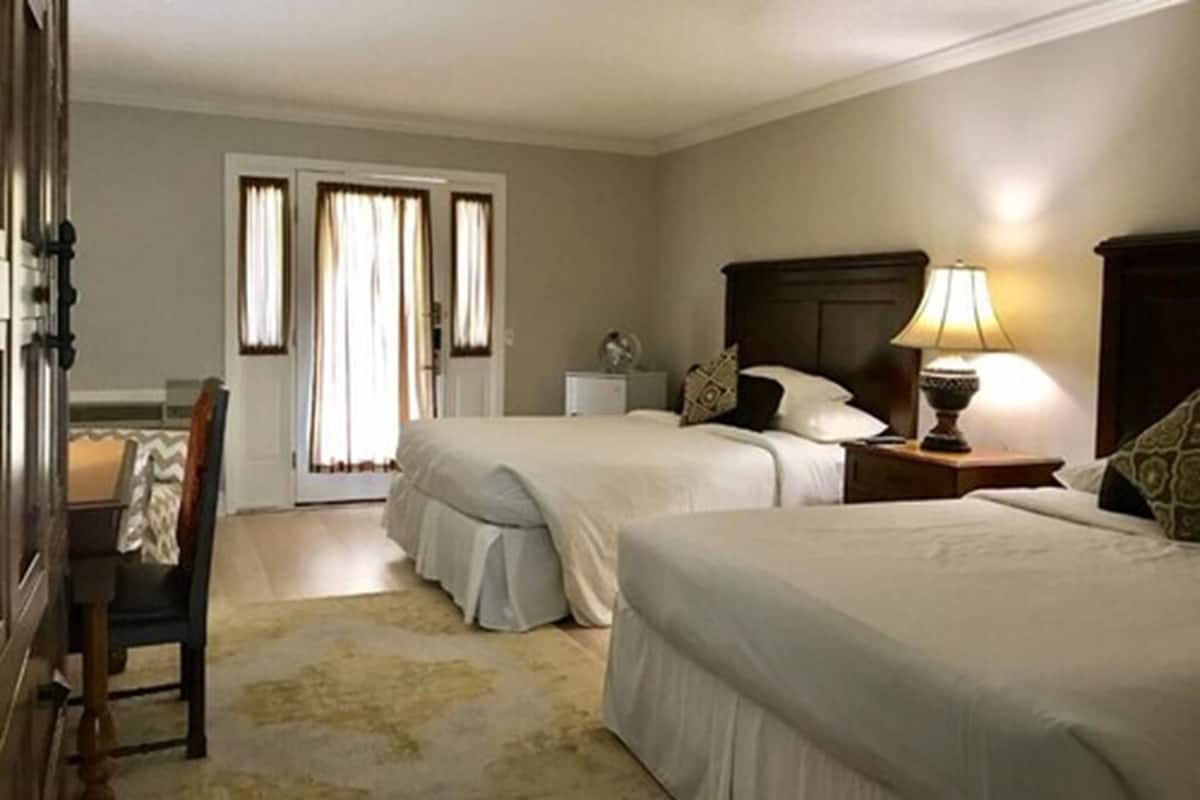 A hotel room with two beds, a bedside table with a lamp, a chair, and a door leading to a balcony.