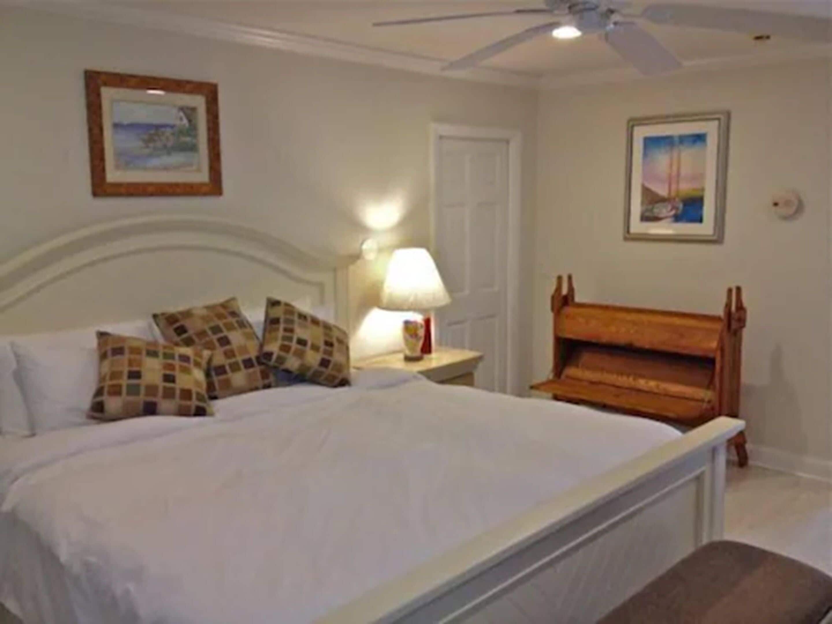 A bedroom with a bed, pillows, a nightstand with a lamp, a wooden bench, and two framed pictures on the wall.