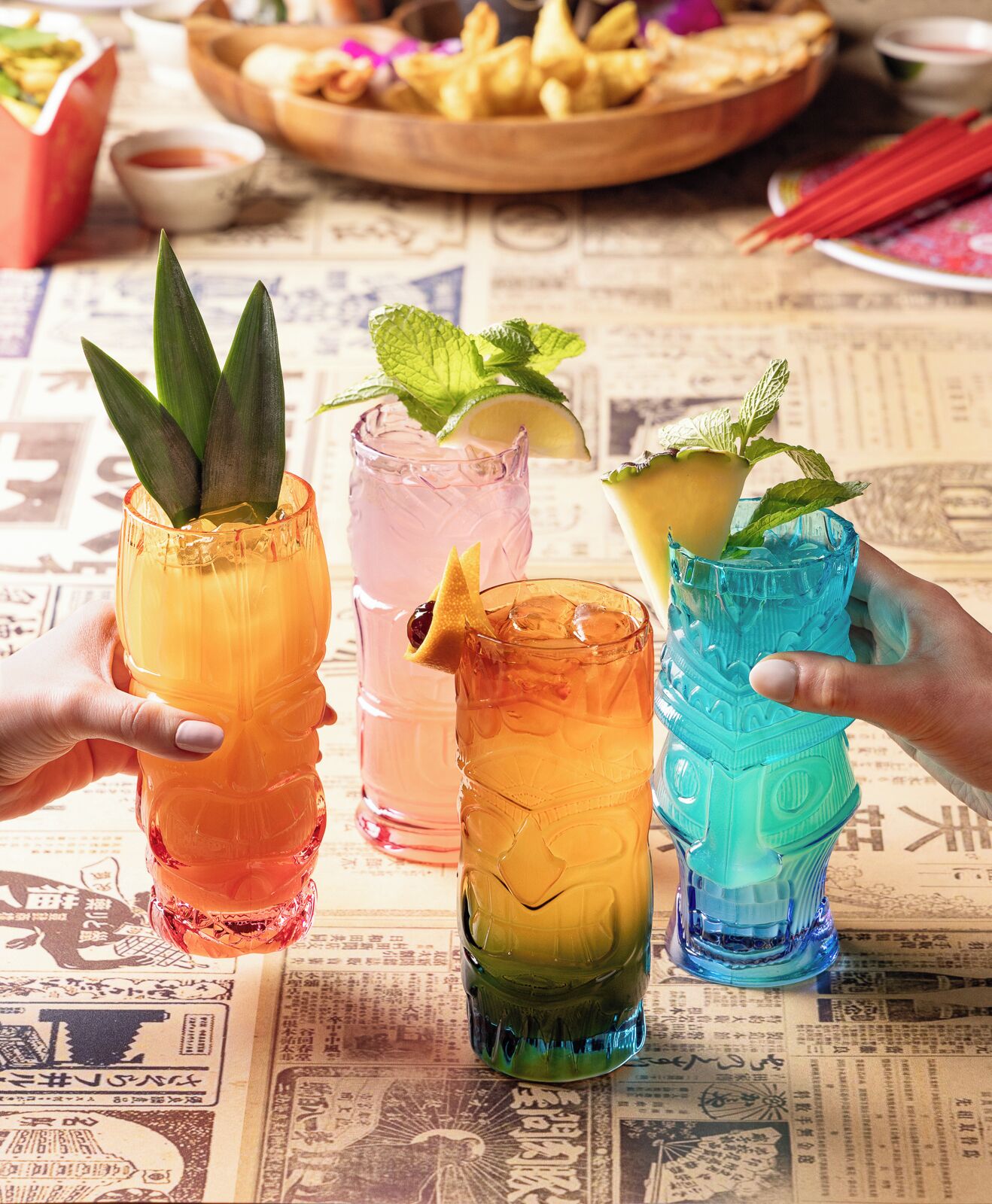 Three colorful drinks with mint leaves, a newspaper-covered table, and a bowl of fries in the background.