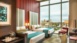 A hotel room with two beds, a desk, a chair, and a large window offering a city view.