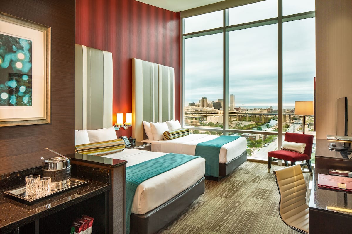Habitación de hotel con dos camas, un escritorio, una silla y un amplio ventanal con vistas a la ciudad.
