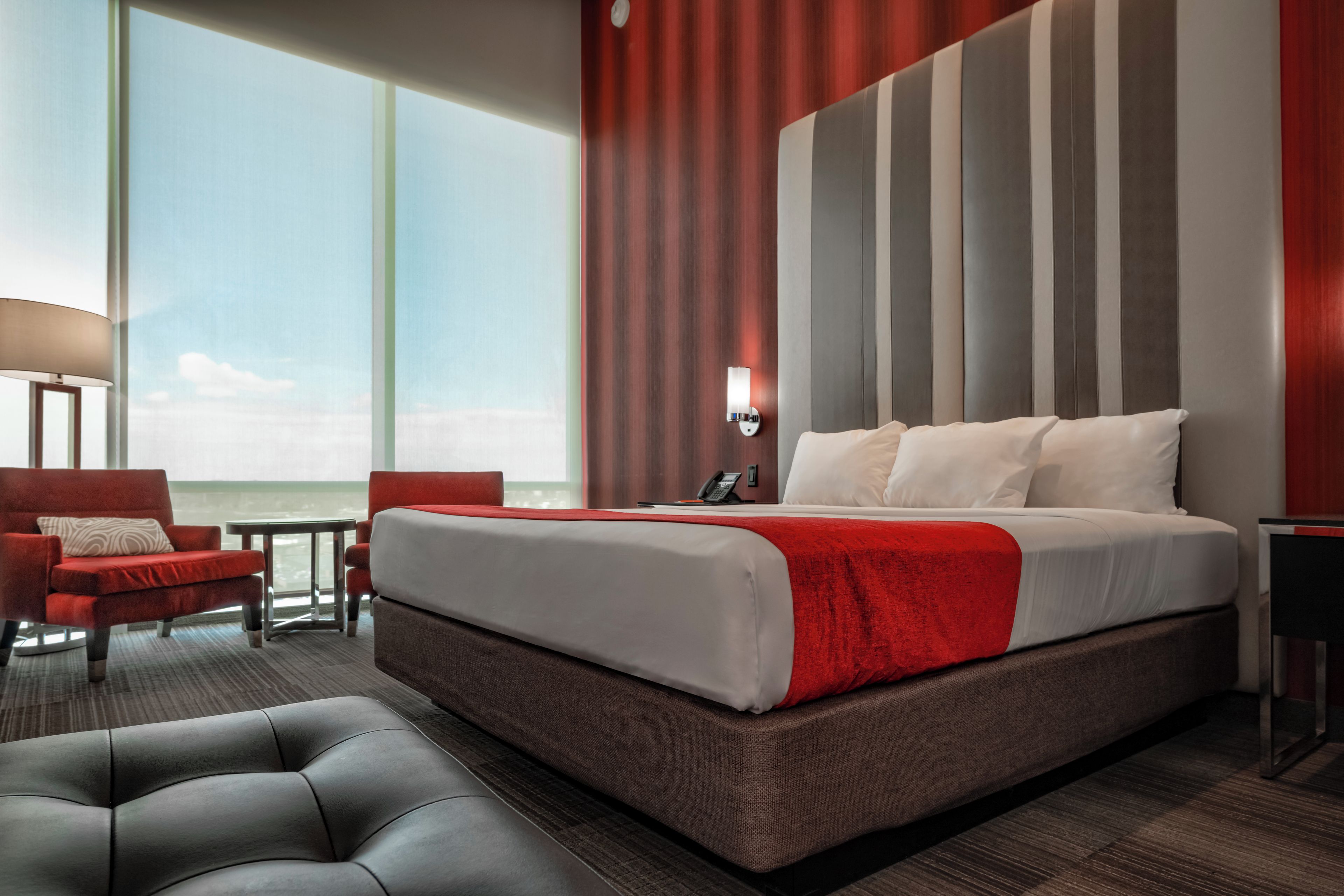 A hotel room with a large bed, a red and white striped headboard, a red armchair, a small table, and a view of the sea.