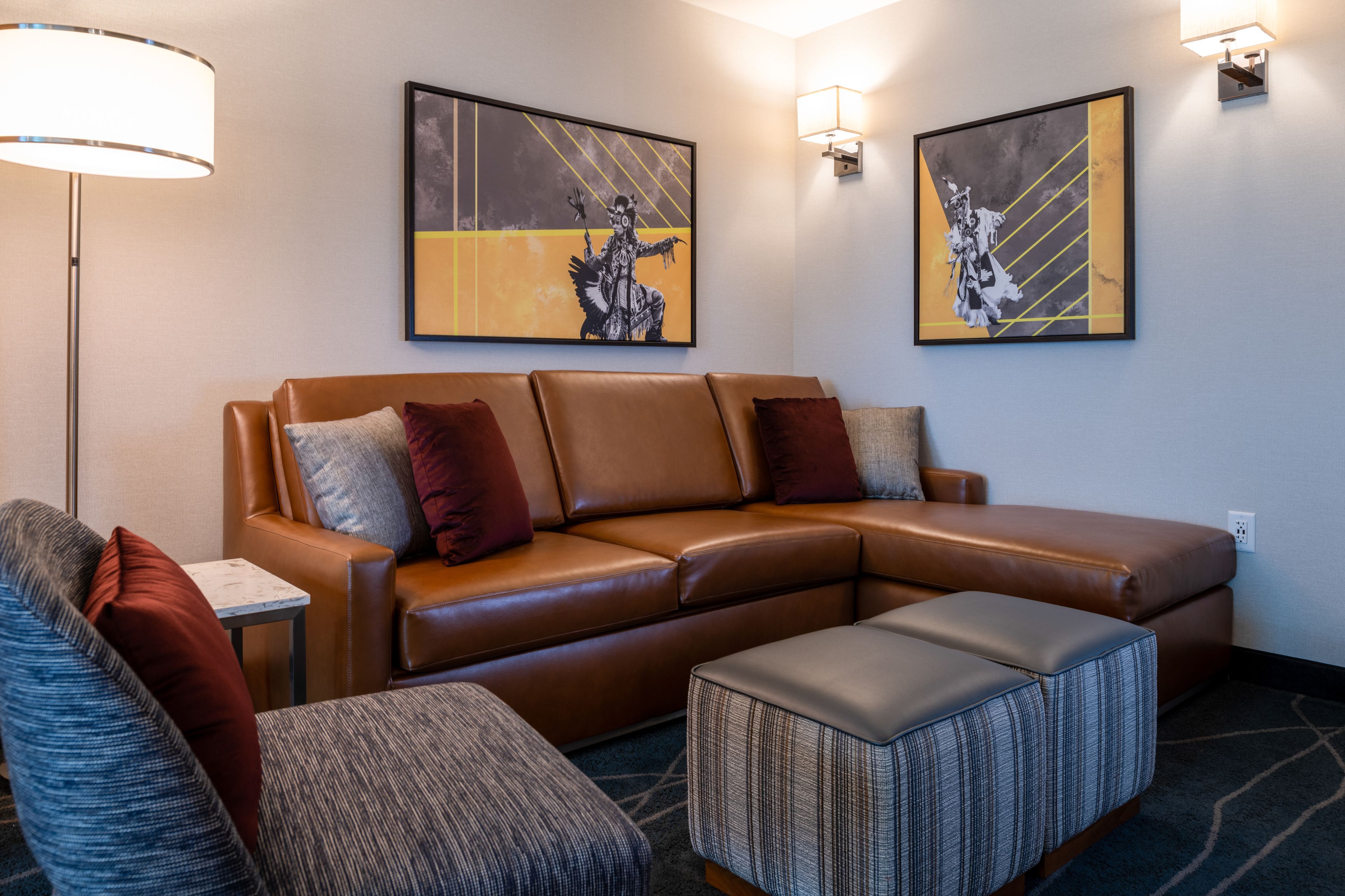 A modern living room with a brown leather sofa, a grey armchair, and a patterned ottoman. There are two wall-mounted artworks depicting abstract figures.