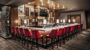Zona de bar con una barra larga, taburetes rojos y una estantería para vinos.