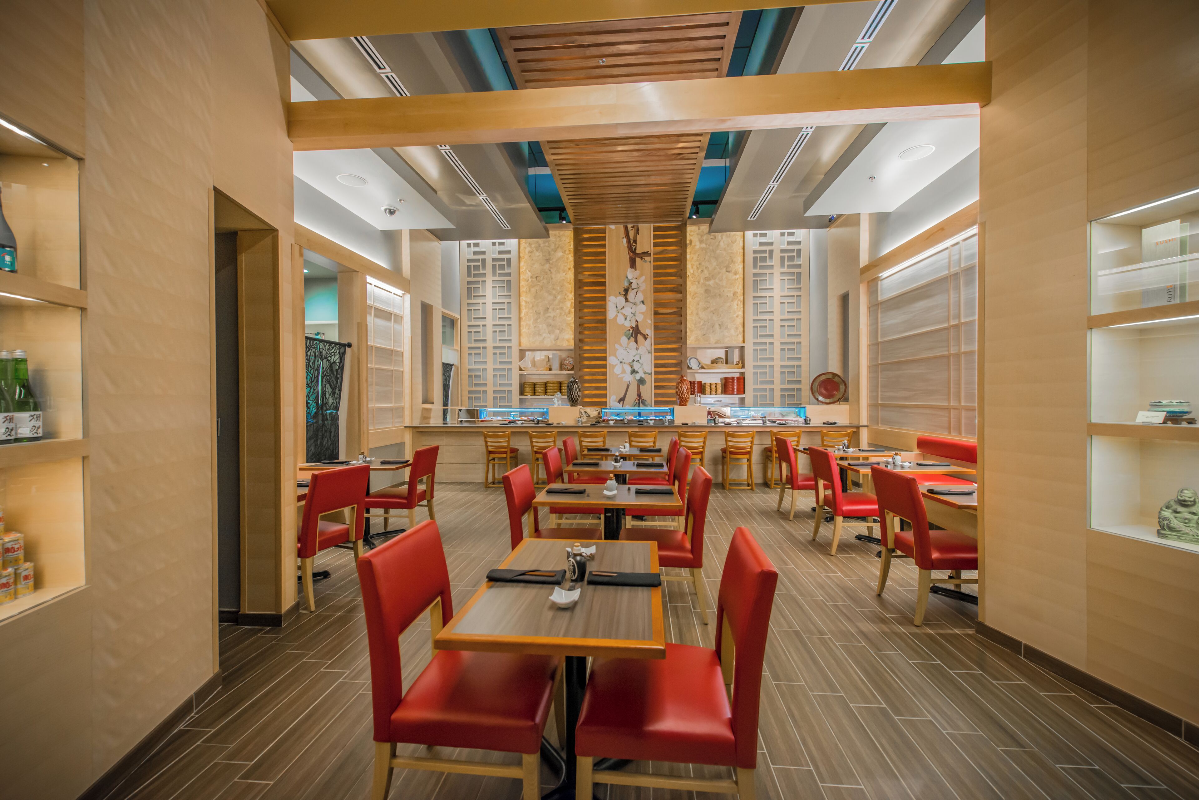 Interior de restaurante con mesas de madera y sillas rojas, una pared decorativa central y estantes con botellas y vajilla.